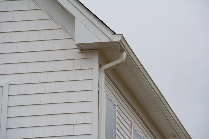 Close-up of the upper side of a house with white siding and trim, featuring a rain gutter system and an overcast sky in the background.