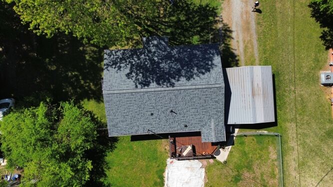 Aerial view of a house with a dark gray roof, covered patio, wooden deck, and trees casting shadows on the lawn.