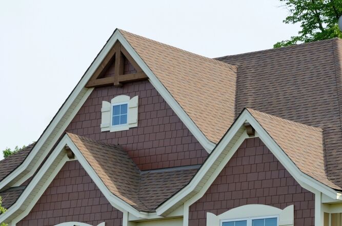 A house roof with multiple gables, brown shingles, and cream trim, featuring a small window with decorative molding near the roof peak.