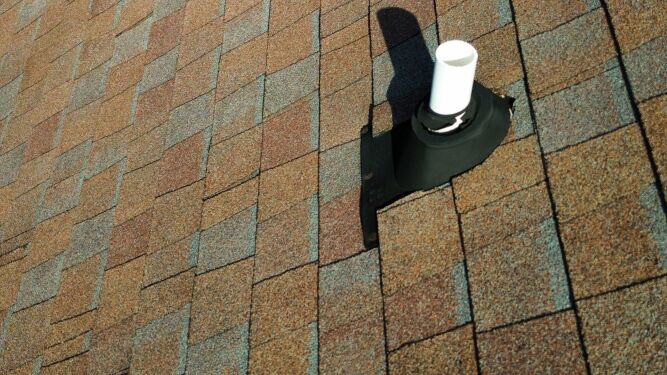 Close-up of a residential roof with asphalt shingles and a white vent pipe surrounded by a black flashing component.