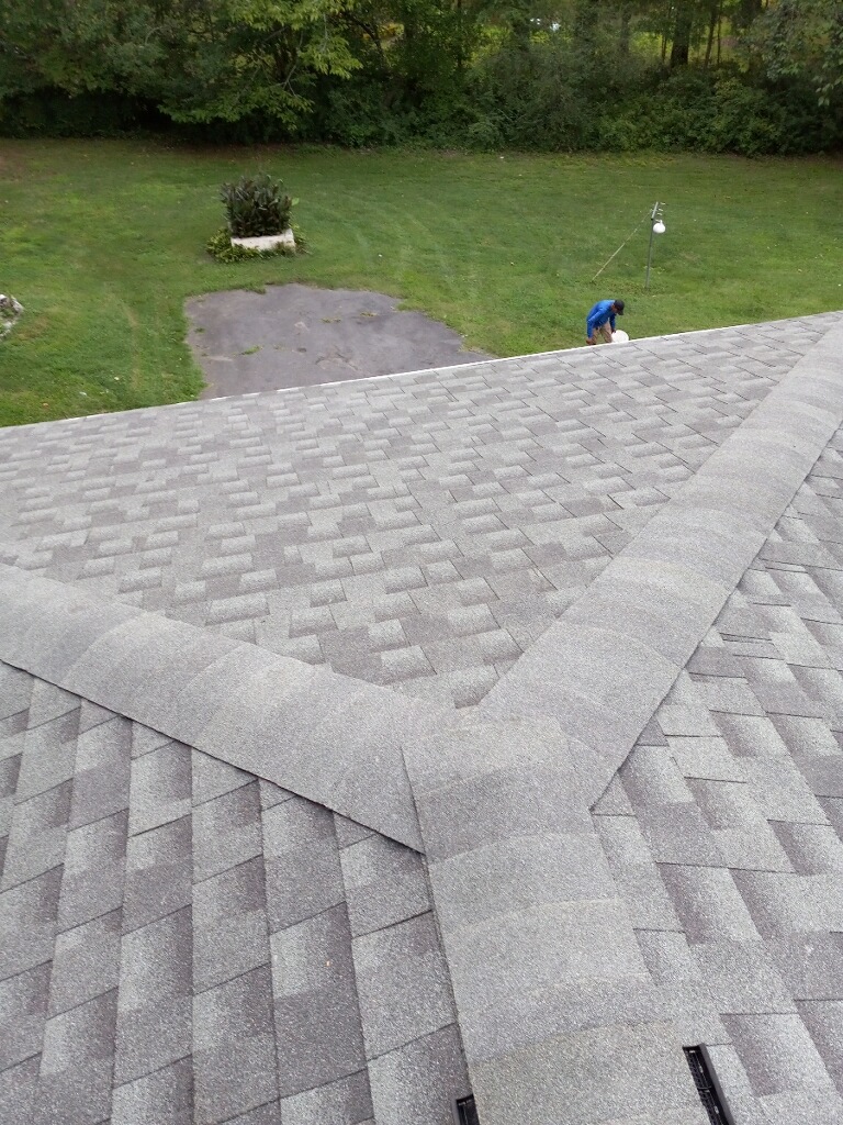 A view from a rooftop with grey shingles, looking down at a person working on the lawn near a paved area, surrounded by green grass and trees.