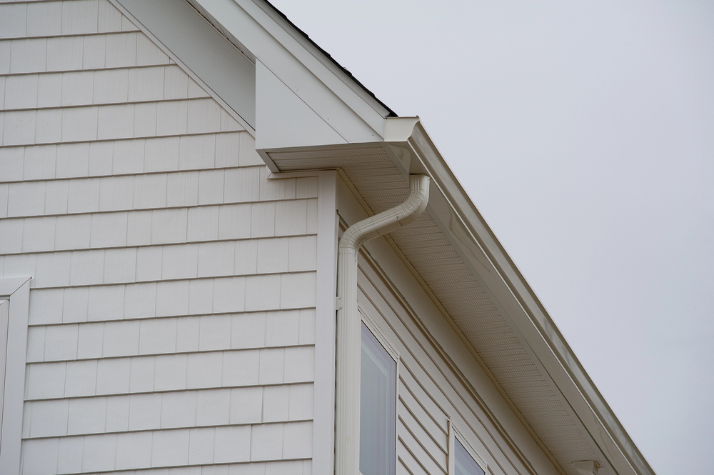 Close-up of the upper side of a house with white siding and trim, featuring a rain gutter system and an overcast sky in the background.