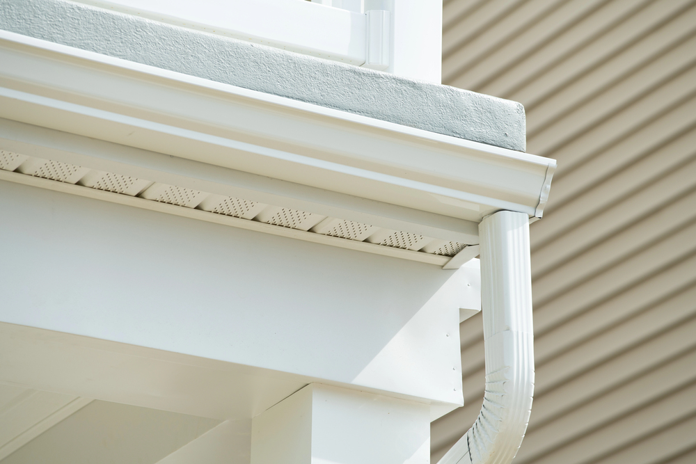 Close-up of white house gutters and downspout attached to a roof with siding in the background.