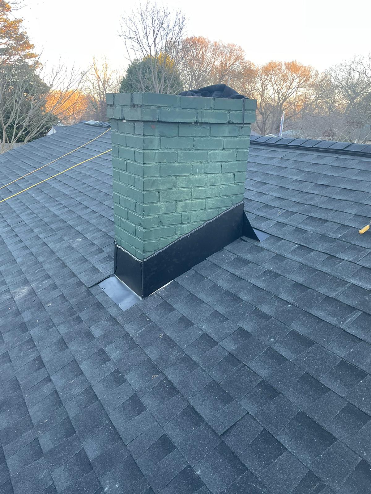 A green brick chimney with black flashing extends from a gray shingled roof; trees without leaves are visible in the background.