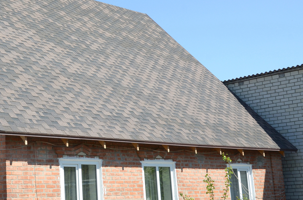 A brick house with a steeply sloped roof covered in gray asphalt shingles, featuring three rectangular windows below the roofline.