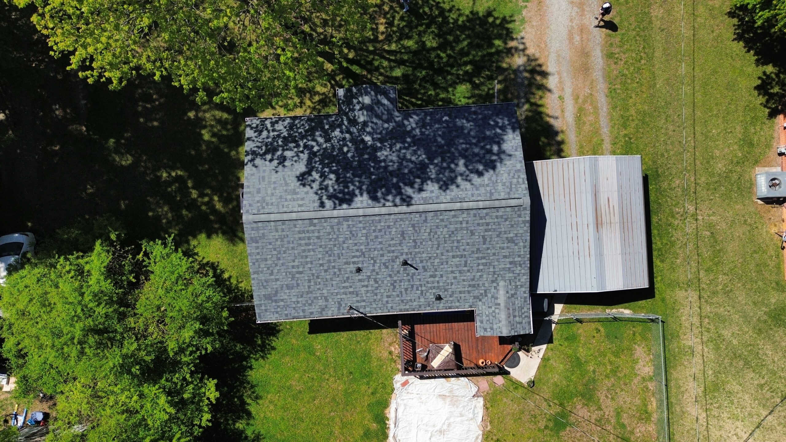 Aerial view of a house with a dark gray roof, covered patio, wooden deck, and trees casting shadows on the lawn.