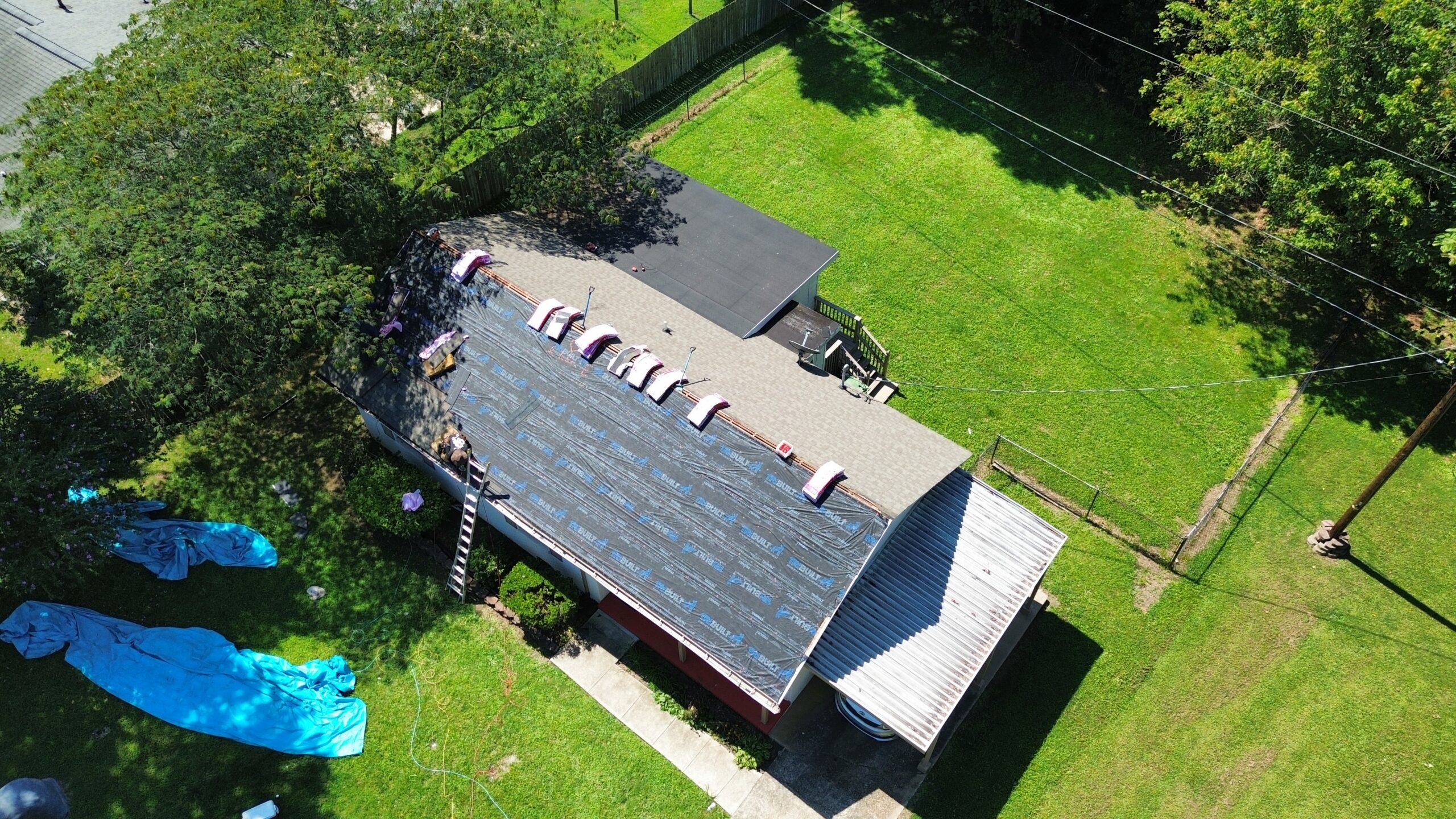 Aerial view of a house roof under construction with roofing materials placed on top and workers visible; surrounding yard is green with trees and tarps on the grass.