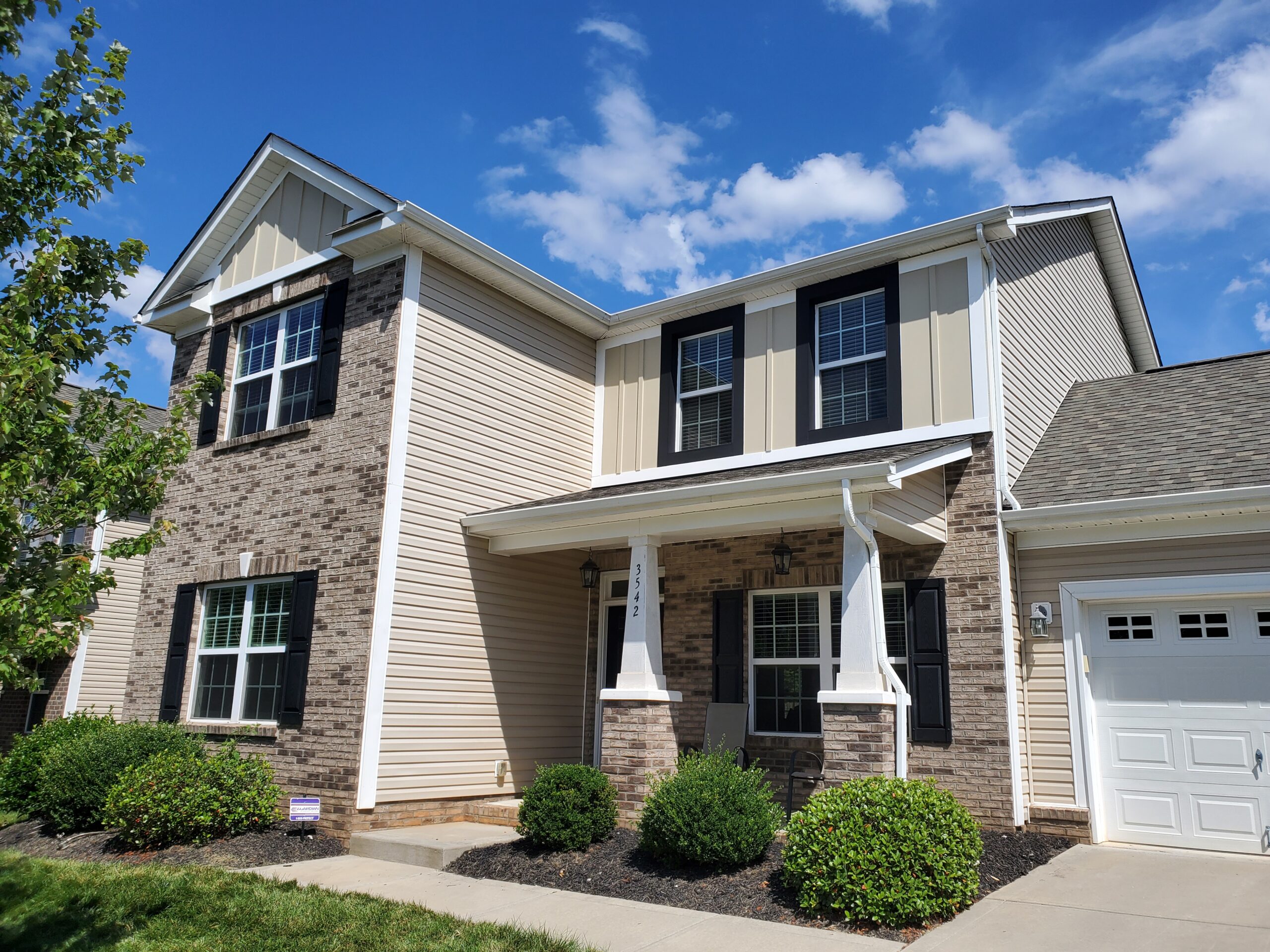 Two-story suburban house with brick and beige siding, black shutters, a small porch, garage, and landscaped front yard under a blue sky with clouds.