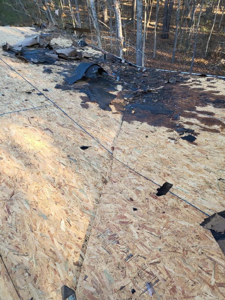 Plywood roof decking with scattered debris and damaged roofing materials near the edge; trees visible in the background.